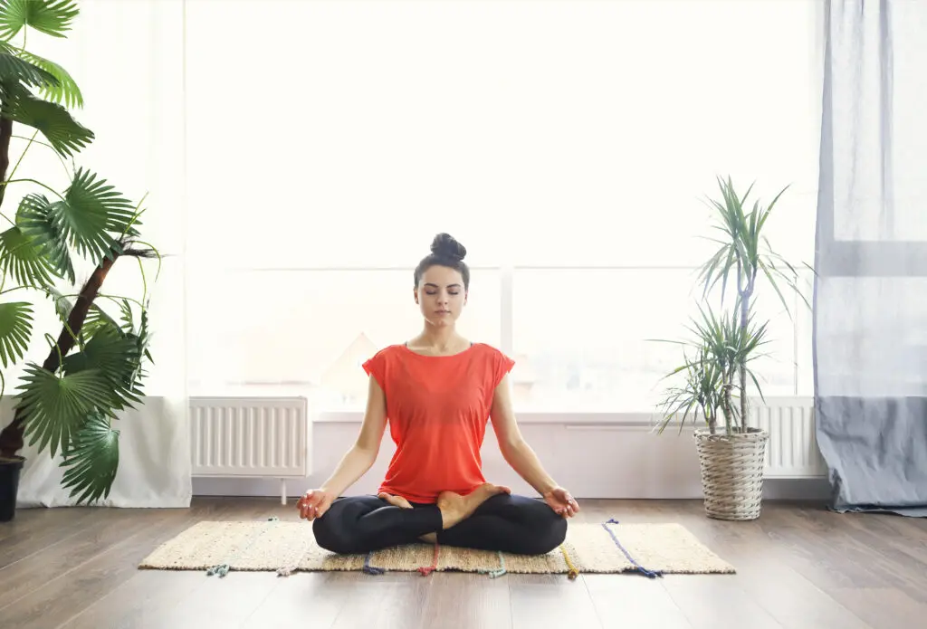 Woman meditating cross-legged on a mat in a bright room.
